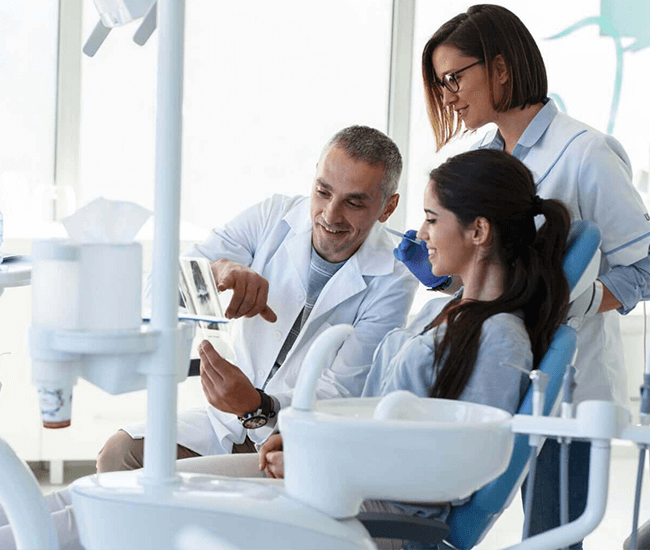 A relaxed and smiling male patient in a blue shirt engages in conversation with his female dentist, who is dressed in a white coat and wearing protective blue gloves and a surgical mask, in a modern and bright dental clinic.