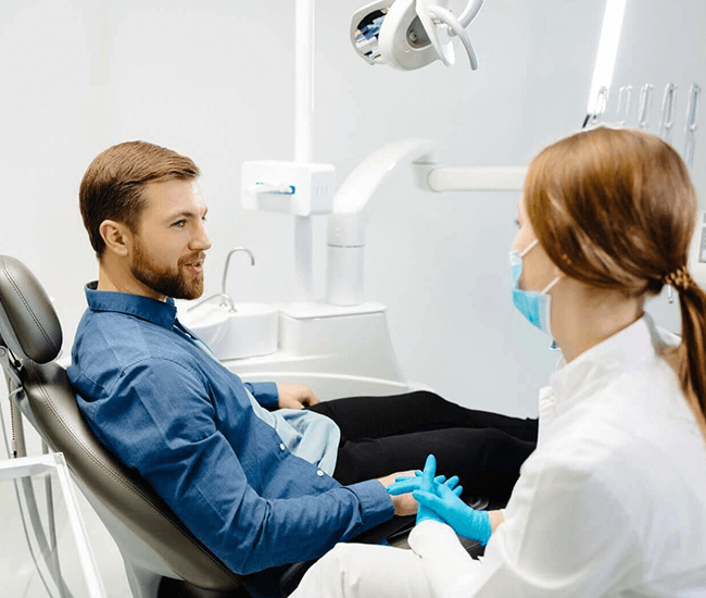 A relaxed and smiling male patient in a blue shirt engages in conversation with his female dentist, who is dressed in a white coat and wearing protective blue gloves and a surgical mask, in a modern and bright dental clinic.